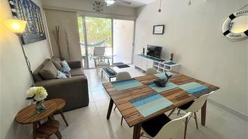 A living room with a sofa, a dining table with chairs, a TV, and a hammock outside the sliding glass door.
