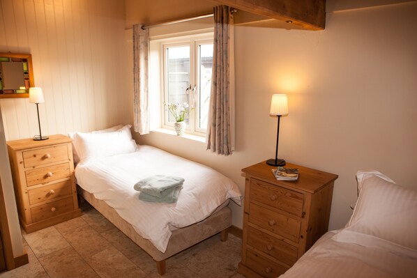 Une chambre avec un lit simple, des tables de chevet en bois, une fenêtre avec des rideaux et une petite plante en pot posée sur le rebord de la fenêtre.
