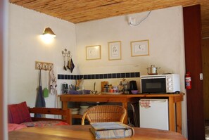A compact kitchen with a microwave, stove, and various utensils. There is a dining area with a chair and a table. - Welcome to #14, a skip & a hop to our famous Muizenberg beach, fun all the way. (Cape Town)