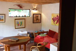 A cozy room with a wooden table, a bed with red bedding, a bookshelf, and a window with a view of greenery. - Welcome to #14, a skip & a hop to our famous Muizenberg beach, fun all the way. (Cape Town)