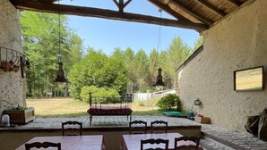 Een terras buiten met stenen vloer en houten tafels en stoelen, overdekt met een houten dak.