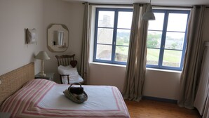 A bedroom with a wooden floor, a bed with a striped bedspread, a bedside lamp, a chair with a heart cushion, a mirror, and a window with curtains.