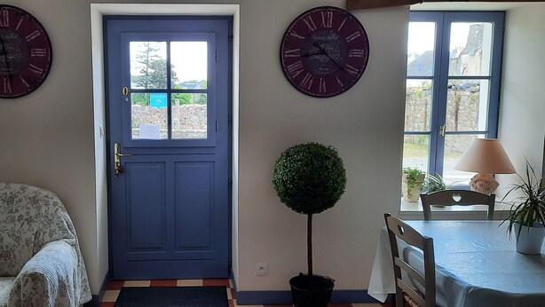 A room with a blue door, two wall clocks, a small round table with chairs, a sofa, and a potted plant.