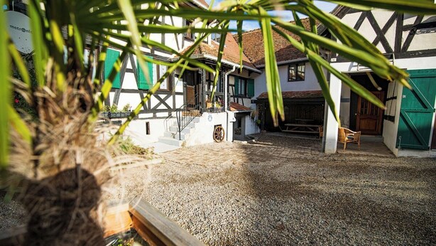 A courtyard with a gravel ground, traditional half-timbered buildings, and a wooden bench. - Les Vignes Vacation Rental - Studio - Route des Vins d'Alsace (Dieffenthal)