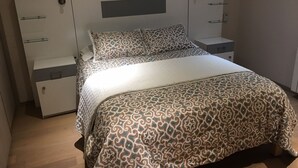 Uma cama bem arrumada com roupa de cama estampada, uma mesinha de cabeceira e um guarda-roupa com gavetas e espaço para pendurar roupas.