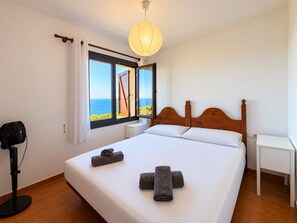 Une chambre d’hôtel avec un lit, des tables de chevet, un ventilateur et une vue sur la mer, visible à travers une grande fenêtre.