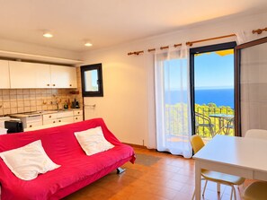 Une pièce avec un canapé rouge, une table blanche, un coin cuisine et un balcon donnant sur la mer.