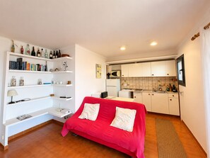 A compact living space with a red sofa, a kitchen area, and a bookshelf filled with books and bottles. - Beautiful studio apartment with spectacular views of the ocean! (Palafrugell)
