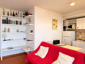 Un espace de vie moderne avec un canapé rouge, des étagères blanches remplies de livres et de bouteilles, un coin cuisine doté d’un four à micro-ondes et une citation inspirante sur le mur.