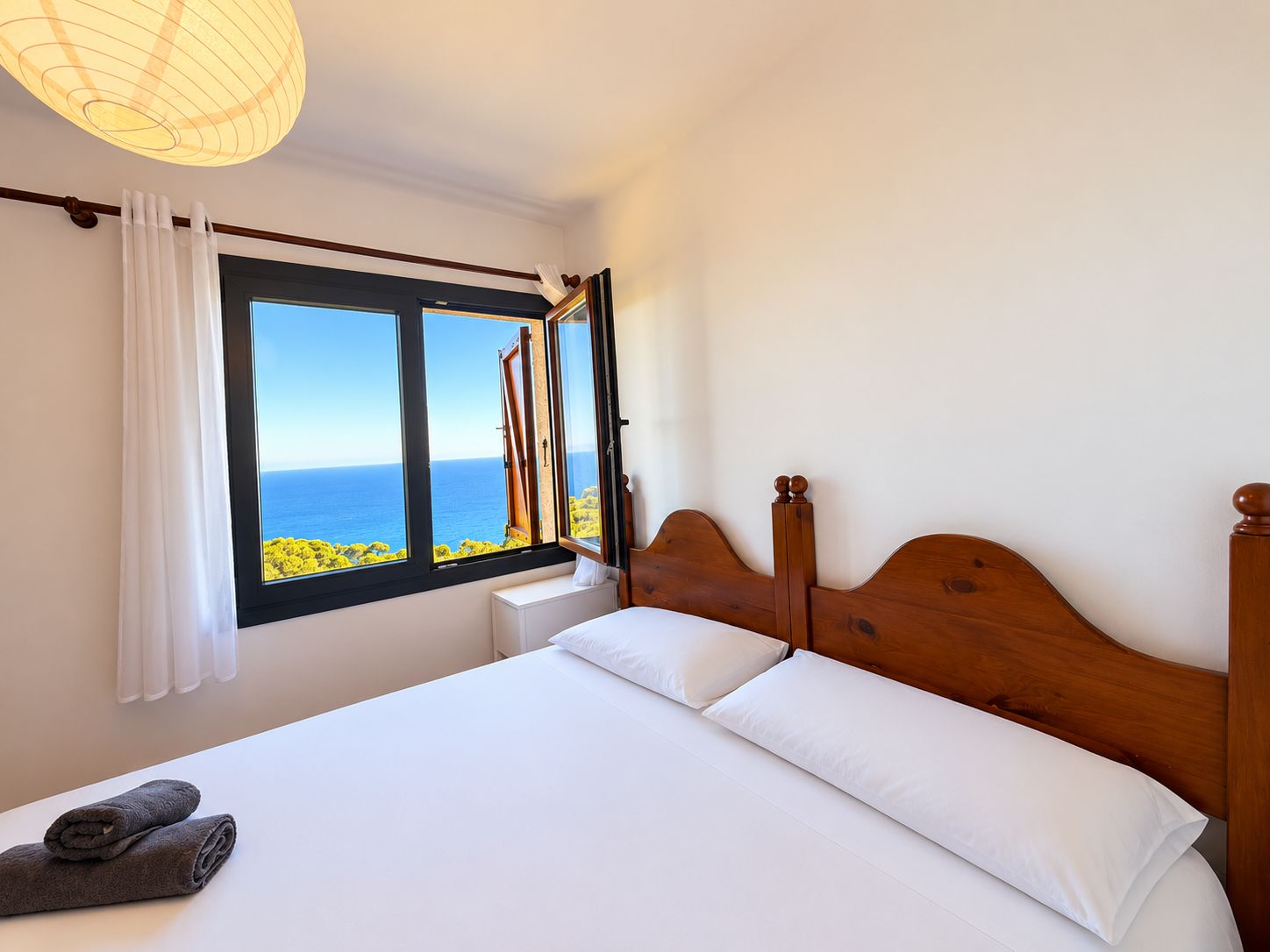 A bedroom with a large window, a wooden bed frame, and a view of the sea.