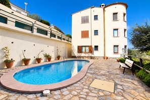 Une piscine entourée d’une terrasse en pierre, agrémentée de plantes en pot, et un bâtiment doté de nombreuses fenêtres et de balcons.