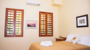 Habitación de hotel con cama, dos ventanas con contraventanas de madera, aire acondicionado, un cuadro enmarcado en la pared y una mesita de noche con lámpara.
