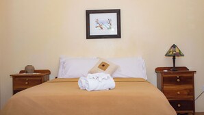 Una cama bien tendida con una manta marrón, una almohada blanca y una toalla enrollada encima. En la pared sobre la cama hay un cuadro enmarcado, una mesita de noche de madera con una lámpara, y un jarrón pequeño en la mesita de noche de la izquierda.