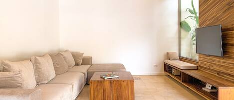 A modern living room with a sectional sofa, a wooden coffee table, a TV mounted on a wooden wall, and a large window with a plant.