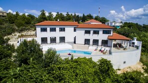 A two-story white building with a red-tiled roof, a swimming pool, and a covered patio area with lounge chairs.