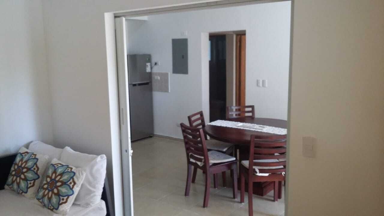 Quarto com mesa de jantar, cadeiras, geladeira e cama com almofadas estampadas.
