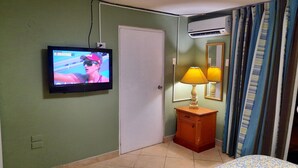 Een kamer met een aan de muur bevestigde televisie, een deur, een klein houten kastje met een lamp en een bed met een bedsprei met dessin.