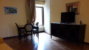 Een woonkamer met een bank, een eettafel met stoelen, een televisie op een kast, en een raam met gordijnen.