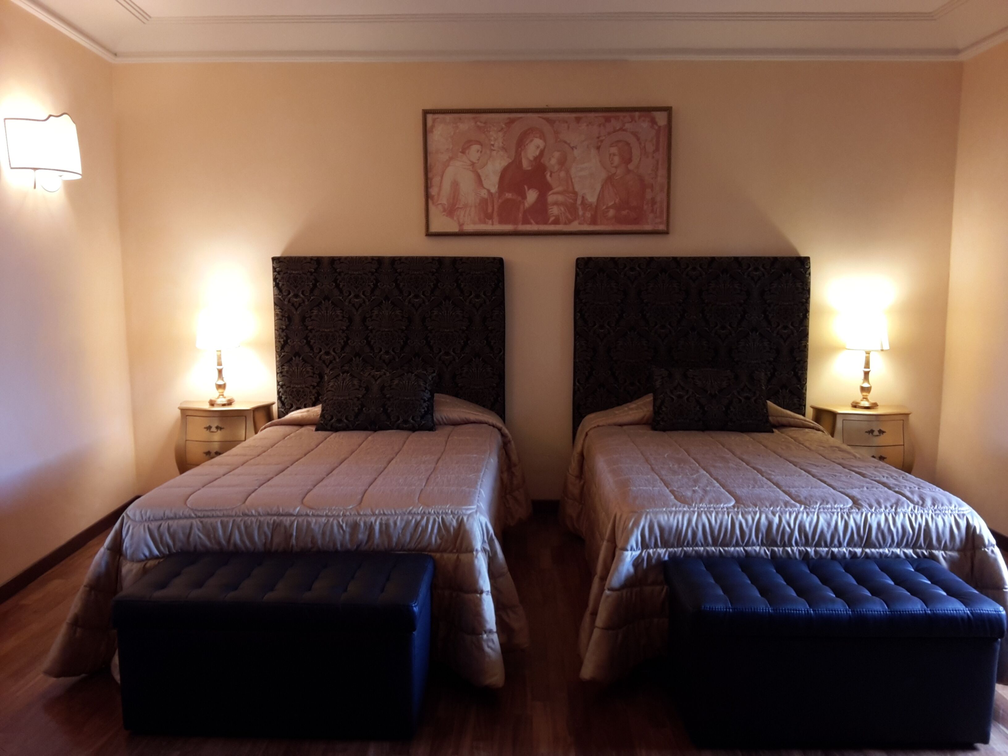 A hotel room with two beds, each with a headboard, bedside tables, and lamps. There is a framed picture on the wall above the beds.