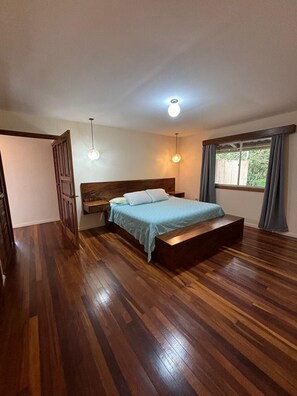 A bedroom with a wooden floor, a bed with a blue bedspread, a nightstand, a hanging light fixture, and a window with curtains.