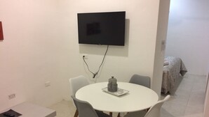 Un pequeño comedor con una mesa redonda, cuatro sillas, un televisor montado en la pared y una cama al fondo.