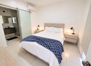 A hotel room with a bed, bedside lamps, a nightstand, a bathroom with a mirror, and an air conditioning unit.