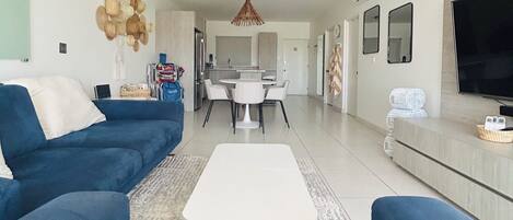 A modern living room with a blue sofa set, a white coffee table, and a dining area with a white table and chairs.