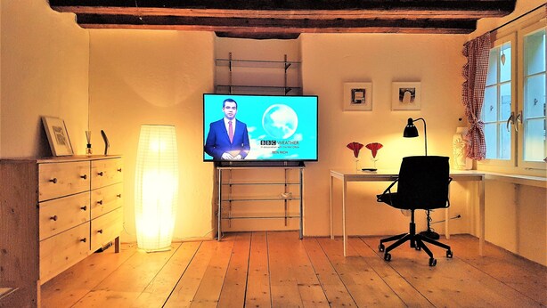 A room with a wooden floor, a desk with a chair, a television displaying a news broadcast, and a window with curtains. - CHALET AU SWISS style (Zürich)