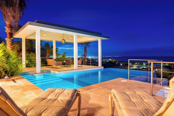 Un espace piscine extérieur moderne, doté d’un coin salon couvert, de chaises longues et offrant une vue sur la ville la nuit.