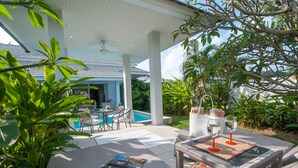 A covered outdoor seating area with a pool, lounge chairs, and a table with magazines and glasses.