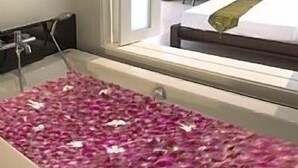 A hotel room with a large bathtub filled with rose petals, a bed with a green blanket, and a view of a cityscape through a large window.