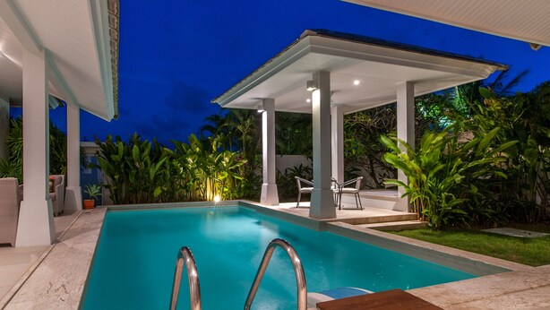 A swimming pool with a covered area, surrounded by greenery and outdoor furniture.