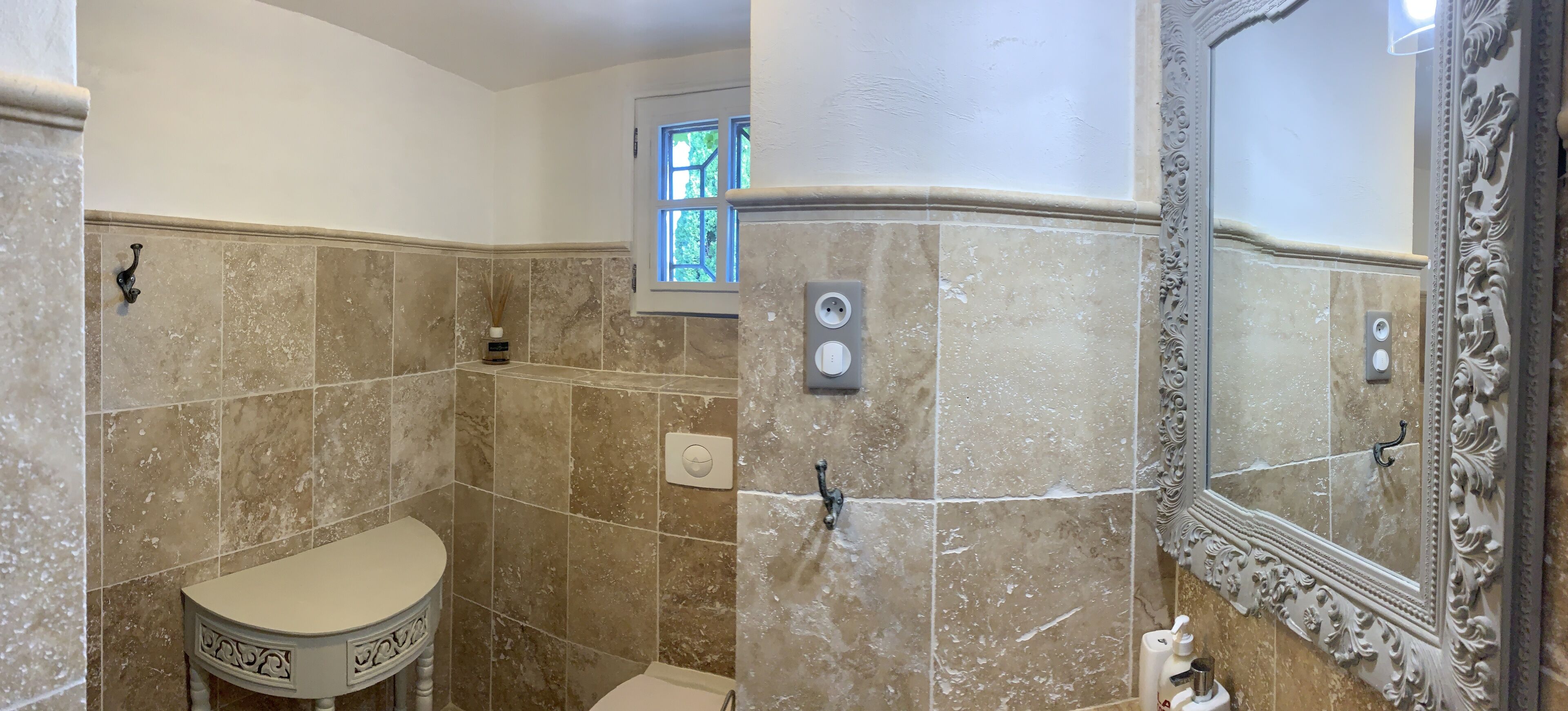 Un baño con baldosas de mármol color crema, un inodoro, un lavamanos, una ducha y un espejo con detalles ornamentales.