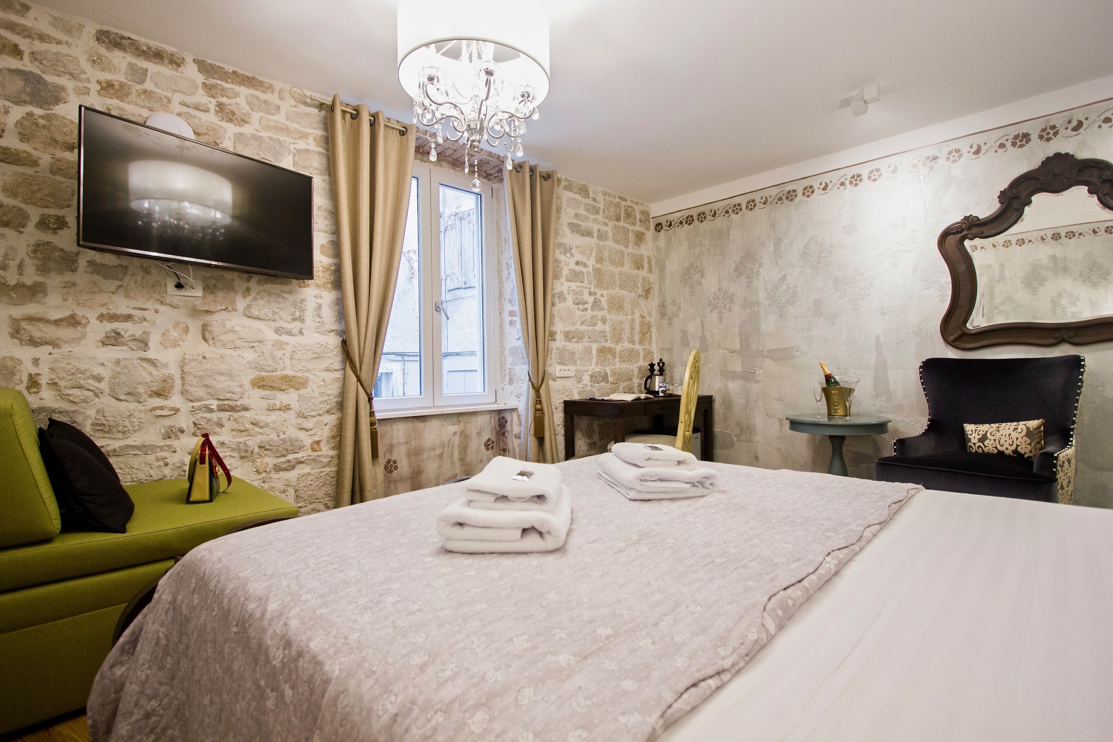 Une chambre d’hôtel avec un mur en pierre, un téléviseur à écran plat fixé au-dessus d’une fenêtre, un lit avec des serviettes soigneusement pliées, un fauteuil vert et une petite table avec une lampe.