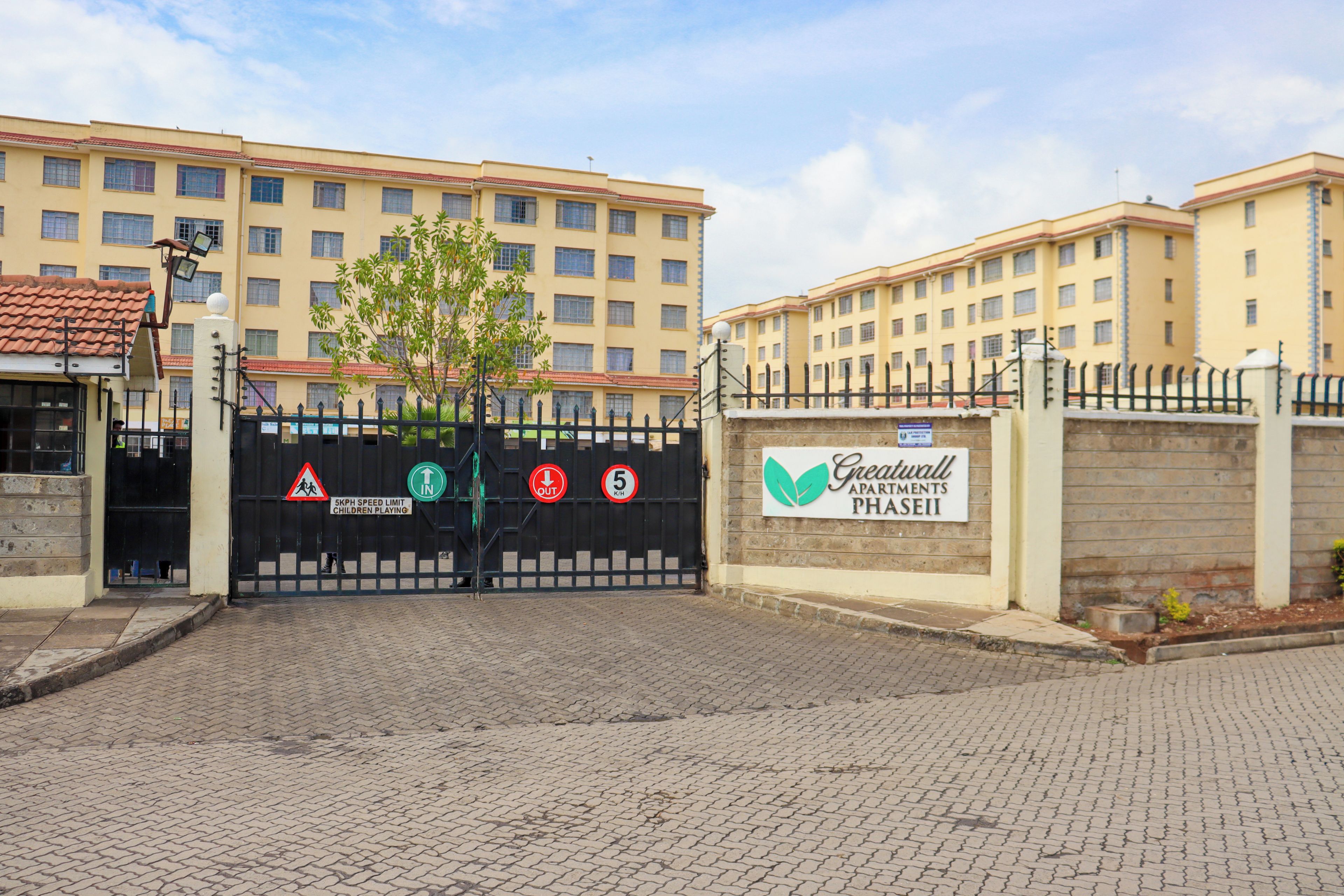A gated entrance to a building with multiple floors, a sign indicating a 5kph speed limit for children playing, and a sign for 'Greatwall Apartments Phase II'.