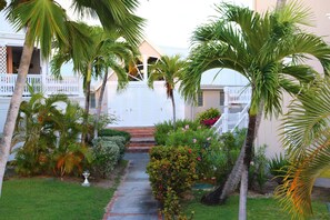 A pathway leading to a white building with a balcony, surrounded by palm trees and various plants. - le "Madras Blue" Duplex, Baie Nettlé, piscine-vue (Marigot)