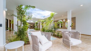 A spacious indoor area with a high ceiling, featuring a central pathway, white wicker chairs, a small table, and various indoor plants. - Luxury Villa: 6 Rooms, 12 Pax, Infinity Pool, Hot Tub, Staff, Close to the Beach (La Romana)