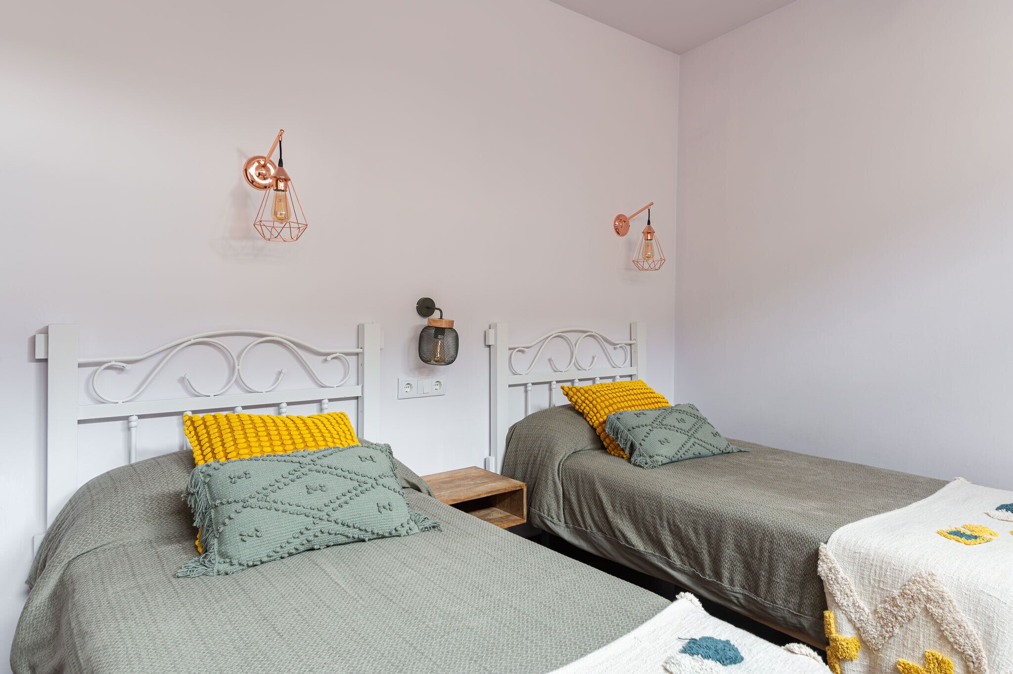 Un dormitorio con dos camas individuales, cada una con una manta gris y un cojín amarillo. Hay dos lámparas fijadas a la pared y una mesita entre las camas.