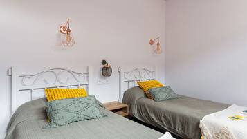 Un dormitorio con dos camas individuales, cada una con una manta gris y un cojín amarillo. Hay dos lámparas fijadas a la pared y una mesita entre las camas.