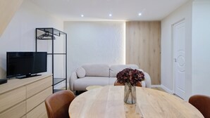 Een moderne kamer met een flatscreen-tv op een houten dressoir, een ronde houten tafel met een bloevas, een beige bank en een kledingrek.