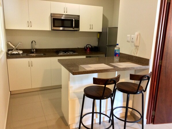 A modern kitchen with white cabinets, a stainless steel microwave, a gas stove, and two bar stools. - Lovely Studio Apartment (Golden Zone) Mazatlan (Mazatlán)