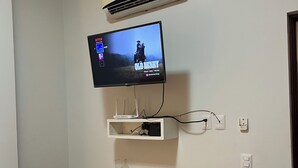 Una habitación con un escritorio de madera, una silla de oficina negra, un televisor montado en la pared mostrando 'Old Henry' y un aire acondicionado instalado en la pared.