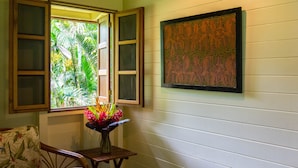 Une pièce avec une fenêtre à cadre en bois, une chaise en bois, une petite table sur laquelle est posé un vase de fleurs et une œuvre d’art accrochée au mur.