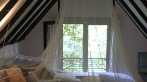 Un dormitorio con cama bajo dosel, ventana con vista a follaje y estructura de techo de madera.