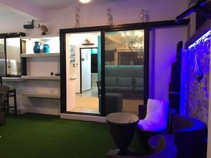 Un salon moderne doté d’une porte vitrée donnant sur un espace repas, d’un petit coin salon extérieur avec un foyer, et d’un mur décoratif agrémenté d’un élément aquatique.