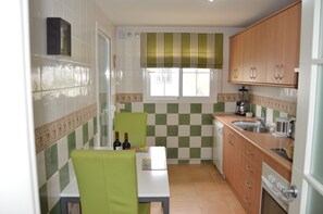 Uma cozinha pequena com painel verde e branco xadrez, uma janela com cortina verde, armários de madeira, uma pia e uma mesa de jantar com cadeiras.