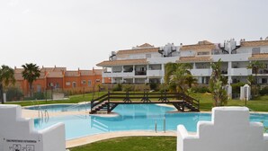 Una piscina con un puente de madera, rodeada por un césped cuidado y palmeras.