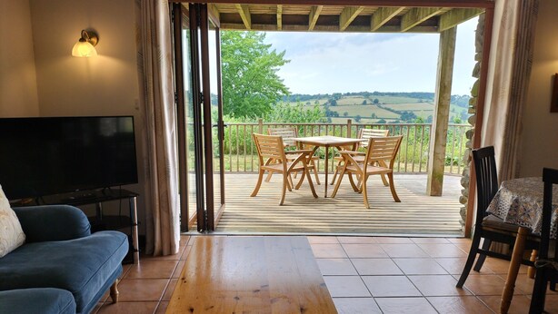 Una sala de estar con televisor, sofá y mesa de madera. Cuenta con un balcón con piso de madera, sillas y vistas al campo.