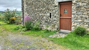 Un edificio de piedra con puerta de madera, rodeado de vegetación y un camino de grava.