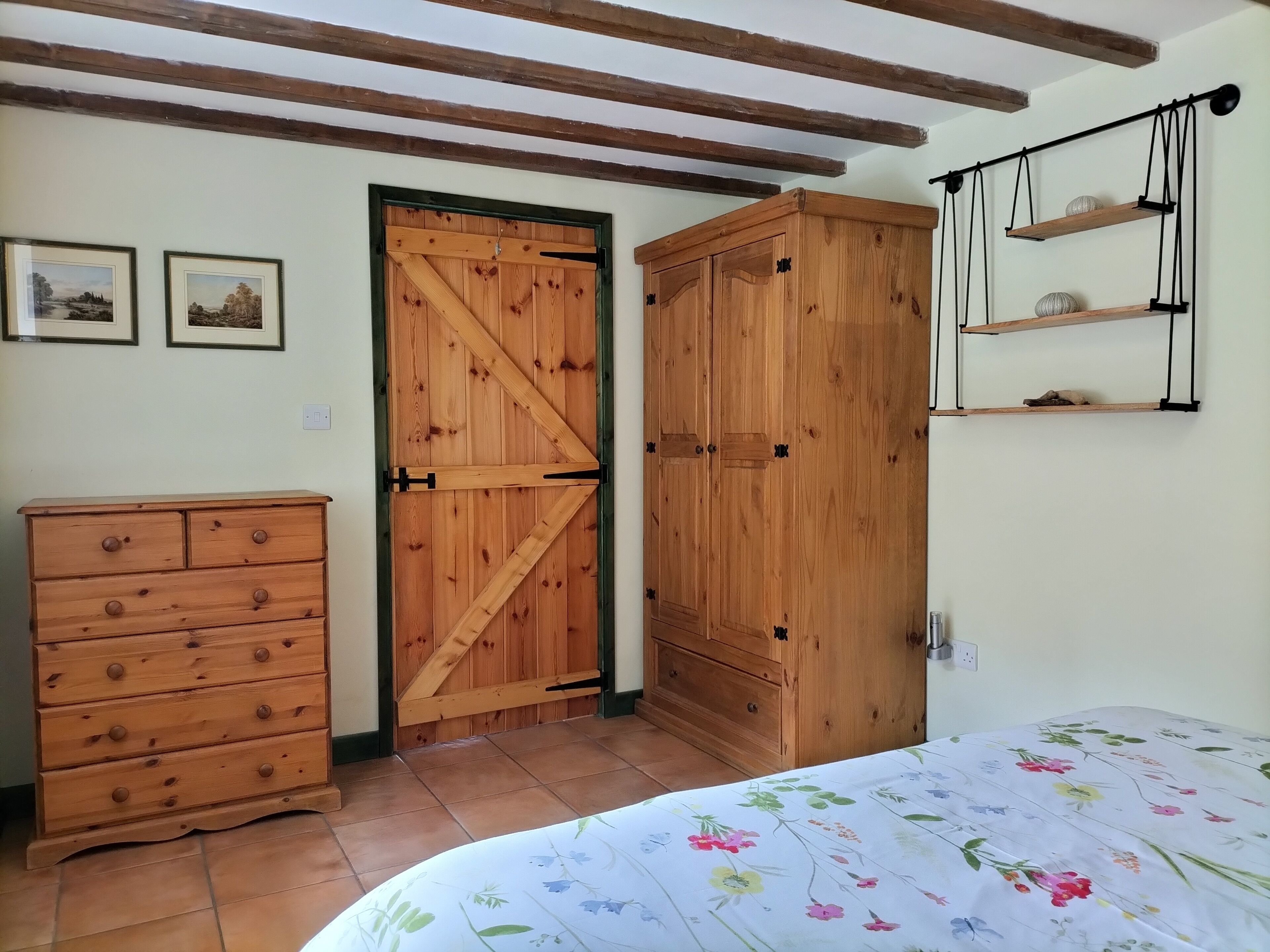Ein Schlafzimmer mit einem Holzschrank, einem Schminktisch, einem Bett mit Blumenbettdecke und einem an der Wand befestigten Regal mit Dekorationen.